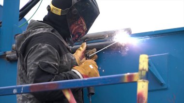 İnşaat alanında mavi metal nesneleri kaynak yaparken koruyucu kaynak maskeli bir adamın yan görüntüsü. Şarjör. İşçi yüksek ısıyı kullanarak metal parçaları eritiyor..