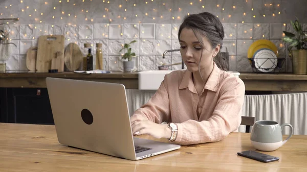 彼女の近代的なアパートの台所で大きなカップと彼女のノートを持つ女性。ストック映像。一般的なラップトップコンピュータの前の台所のテーブルに座っている深刻な若い女性. — ストック写真