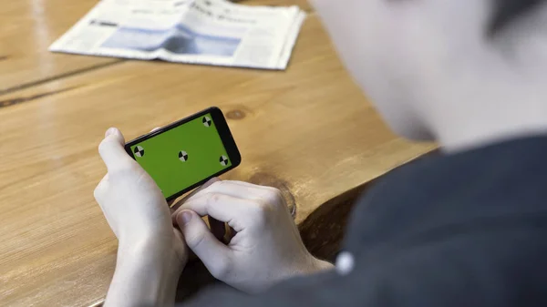 Orientación horizontal del smartphone con pantalla verde: disparo sobre el hombro de un joven. Imágenes de archivo. Hombre sentado en la mesa, tocando y desplazándose en el cromakey del teléfono . — Foto de Stock