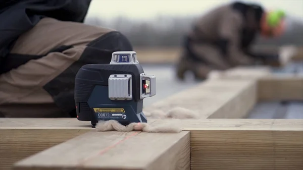 Een arbeider in speciaal beschermend uniform nivelleert een houten balk terwijl hij een fundering bouwt. Een knip. Sluiten voor een spirit laser lavel gereedschap staand op een houten staaf. — Stockfoto