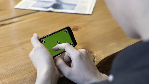 Un tipo sosteniendo en brazos gadget electrónico con cromakey pantalla verde, desplazándose y golpeando sobre fondo de mesa de madera. Imágenes de archivo. Humano sentarse y utilizar el teléfono móvil de mano en la habitación . — Foto de Stock