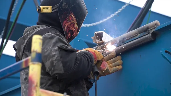 Side view of the worker in fireproof uniform welding parts of blue colored metal construction. Clip. The man in welding mask welding details with many flying sparks.