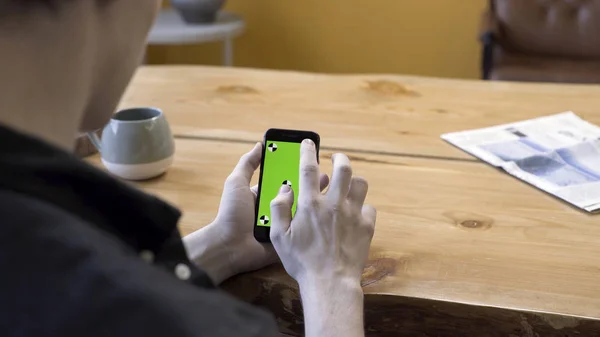 One guy holding in arms electronics gadget with green screen chromakey, scrolling and tapping it on wooden table background. Stock footage. Human sit and use the handheld cellphone in room.