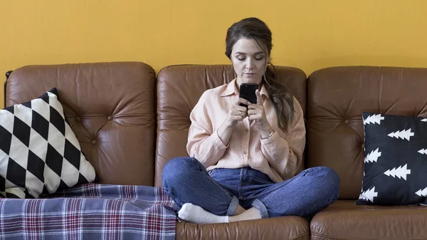 Όμορφη νεαρή γυναίκα με το ανοιχτό ροζ πουκάμισο κρατώντας ένα smartphone στα χέρια της. Στικ. Ενοχλημένη μελαχρινή δακτυλογράφηση και κύλιση iphone ενώ συνομιλούν στο σαλόνι στο σπίτι. — Φωτογραφία Αρχείου