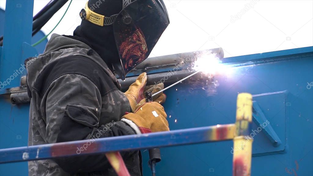 Side view of a man in protective welding mask while welding blue metal objects process at the construction site. Clip. Worker using high heat to melt the metal parts together.
