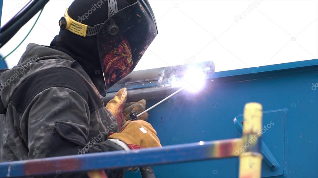 Side view of a man in protective welding mask while welding blue metal objects process at the construction site. Clip. Worker using high heat to melt the metal parts together.