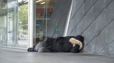 Homeless man in warm clothes sleeping outdoors near the entrance of the building, poverty concept. Stock footage. Poor unemployed male tramp lying near the concrete wall. clipart