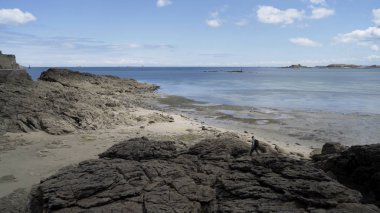 Sırt çantası takan genç bir adam mavi bulutlu arka planda, güzel denizin kıyısında, taşlı sahilde yürüyor. Başla. Uçsuz bucaksız deniz manzarası ve seyahat eden adamla siyah kayalar..