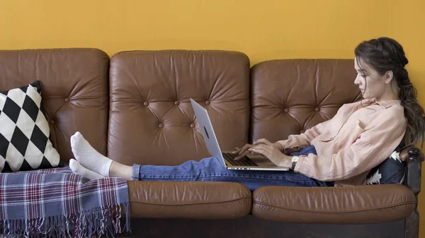 Seitenansicht der attraktiven jungen brünetten Frau, die am Laptop arbeitet und lächelt, während sie auf der Couch im stilvollen Zimmer sitzt, freiberufliches Konzept. Archivmaterial. Weibchen benutzt ihren Laptop zu Hause. — Stockfoto
