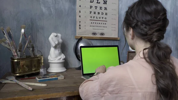 Over the shoulder shot of a woman typing on a computer laptop with a chromakey green screen. Stock footage. Woman hands typing on laptop with green screen in the artist workshop.