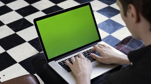 Over the shoulder view of a man using his computer with green screen and sitting on a couch, time lapse. Stock footage. Businessman typing on his laptop with chromakey on checkered floor background.