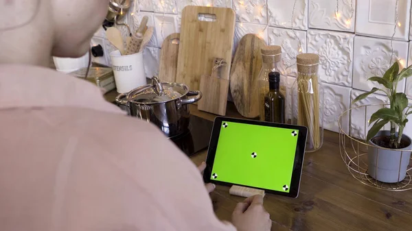 Rear view of a woman tapping on a tablet with green screen in the kitchen at home for recipes. Stock footage. Female using her tablet with chroma key for cooking.