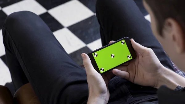Business man using smart phone with green screen while sitting on a couch on checkered black and white floor background. Stock footage. Man holding device in horizontal position and watching. — Stock Photo, Image