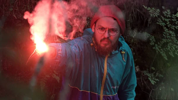 Man lost in the forest holding red burning signal flare to draw attention. Stock footage. Male wearing blue jacket and red hat swinging his hand with a signal flare in the woods.