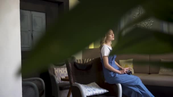 Hermosa chica rubia con camiseta blanca y mono azul sentado en la sala de estar y la celebración de la cámara, vista desde detrás de las hojas de la planta verde. Art. Mujer joven reflexiva dentro del apartamento . — Vídeos de Stock