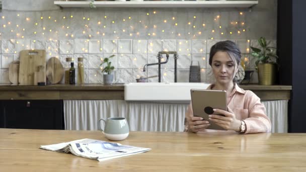 Belle jeune femme utilisant une tablette numérique dans la cuisine avec un mur décoré d'une guirlande sur le fond. Images d'archives. Femme assise à la table en bois avec une tasse à thé et un journal . — Video