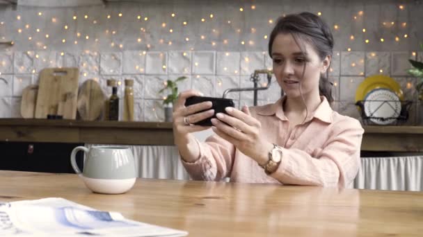 Un retrato de una joven morena feliz con camisa rosa claro y reloj de oro jugando juegos en el teléfono móvil en su cocina. Imágenes de archivo. Gadget moderno en manos femeninas . — Vídeos de Stock