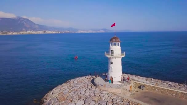 Vista superior del faro en el fondo del mar y la ciudad turística. Clip. Hermoso faro blanco se encuentra en el mar azul con vistas a las montañas con la ciudad en el horizonte — Vídeo de stock