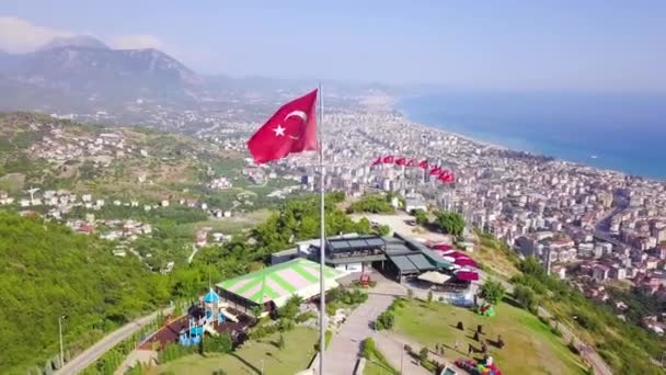 Draufsicht der türkischen Flaggen auf dem Berg vor dem Hintergrund der Stadt und des Meeres. Clip. türkischer Ferienort in bergiger Gegend am Strand — Stockvideo