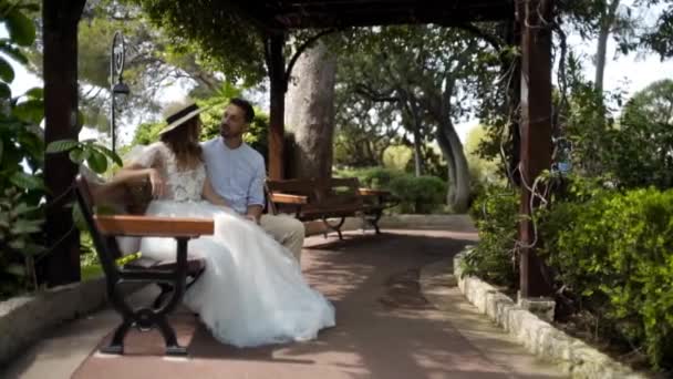 Recién casados sentados en el banco en el jardín verde. Acción. Hermosa pareja joven están sentados y charlando en el banco en un hermoso cenador de exuberante jardín — Vídeos de Stock