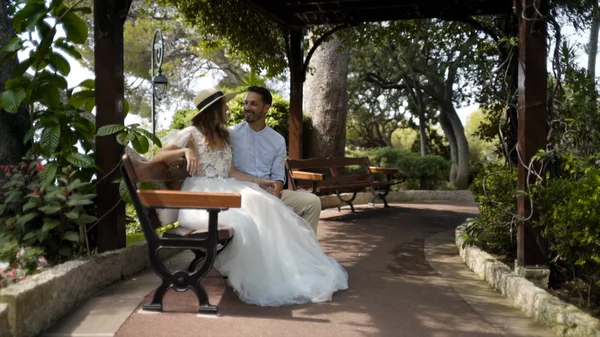 Frischvermählte sitzen auf einer Bank im grünen Garten. Aktion. schönes junges Paar sitzt und plaudert auf Bank in schöner Laube des üppigen Gartens — Stockfoto