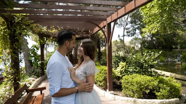 Frischvermählte schauen sich vor dem Hintergrund des Gartens liebevoll an. Aktion. Schönes junges Paar schaut einander liebevoll im sonnigen, grünen Garten stehend an — Stockfoto