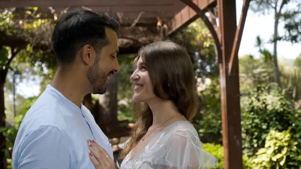 Newlyweds lovingly look at each other on background of garden. Action. Beautiful young couple lovingly look at each other standing in sunny green garden