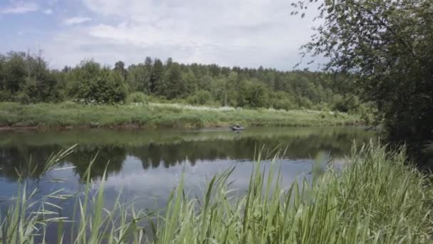 Piękny letni krajobraz z rzeką i jej zielonymi brzegami i człowiekiem pływającym w nadmuchiwanej łodzi. Materiał filmowy. Rybak na nadmuchiwanej gumowej łodzi na rzece. — Wideo stockowe