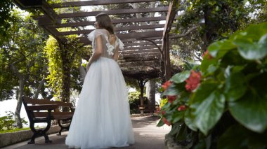 Genç güzel gelin bahçede beyaz elbise standı. Eylem. Gazebo altında yeşil bahçede duran narin beyaz elbiseli genç gelinin arka görünümü