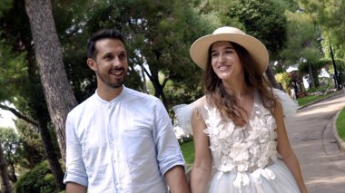 Yeni evliler gülümsüyor ve parkta el ele tutuşuyorlar. Eylem. Güzel genç çift balayı mutluluk ile ışınlanıyor. Şapkalı genç kadın kocasına gülümsüyor