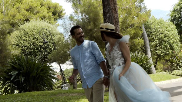 Newlyweds run in park in summer. Action. Beautiful loving couple romantically run away holding hands in park. Wife in white dress holding her husbands hand illuminated by sunlight — Stock Photo, Image