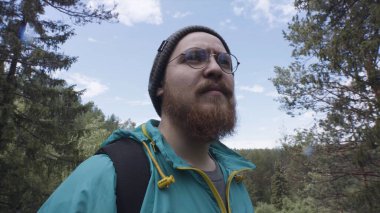 Hippi yürüyüşçünün mavi bulutlu arka plandaki yeşil vadiyi gözlemlediği yakın plan. Stok görüntüleri. Güzel bir ormana bakan yuvarlak gözlüklü sakallı bir adam..