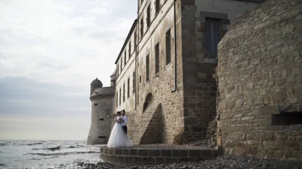 Happy romantic wedding couple standing by the sea near the old castle, photosession outdoors, love and family concept. Action. Bride and groom on the sea shore on their wedding day. — Stock Photo, Image