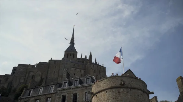 Widok dolny zamku Mont Saint Michel we Francji na tle błękitnego zachmurzonego nieba z latającymi mewami. Akcja. Piękny stary zabytkowy budynek otoczony przez latające ptaki. — Zdjęcie stockowe