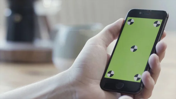 Close up of hands using smartphone with green screen, sliding and tapping on blurred kitchen table background. Stock footage. Man hands holding mobile phone with chroma key.