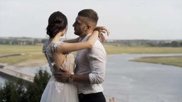 Stilvolle Braut und Bräutigam posiert vor dem Hintergrund des Flusses und der grünen Wiese, Liebe und Familienkonzept. Aktion. Sinnliches Hochzeitspaar umarmt und teilt die Liebe. — Stockfoto