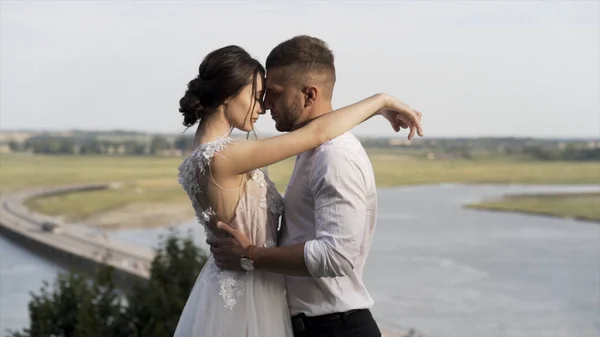 Stilvolle Braut und Bräutigam posiert vor dem Hintergrund des Flusses und der grünen Wiese, Liebe und Familienkonzept. Aktion. Sinnliches Hochzeitspaar umarmt und teilt die Liebe. — Stockfoto