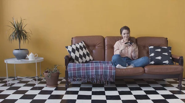 Feliz joven y hermosa mujer jugando un juego en su teléfono inteligente, sentado en un sofá en casa en la habitación con estilo brillante. Imágenes de archivo. Mujer divirtiéndose, jugando su dispositivo . —  Fotos de Stock