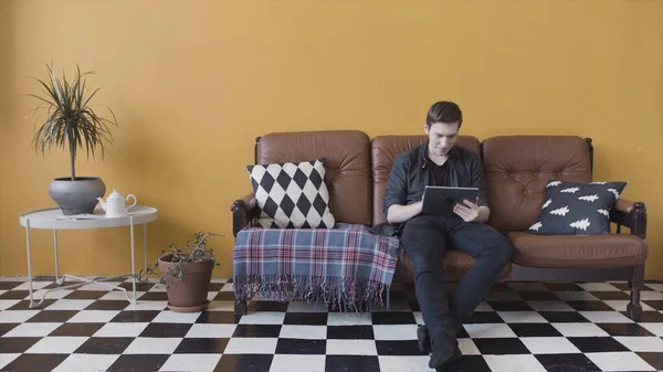 Красивий випадковий молодий чоловік, використовуючи планшет вдома, сидить на дивані у своїй вітальні, переглядаючи Інтернет. Стокові кадри. Серйозна людина використовує свій пристрій у світлій незвичайній кімнаті . — стокове фото