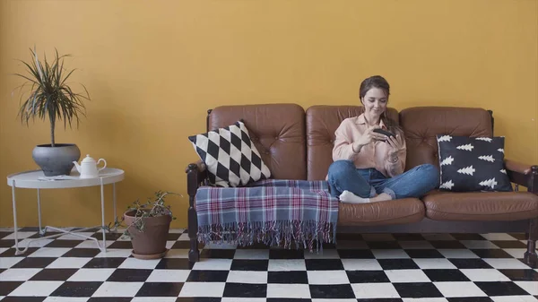 Feliz joven y hermosa mujer jugando un juego en su teléfono inteligente, sentado en un sofá en casa en la habitación con estilo brillante. Imágenes de archivo. Mujer divirtiéndose, jugando su dispositivo . —  Fotos de Stock