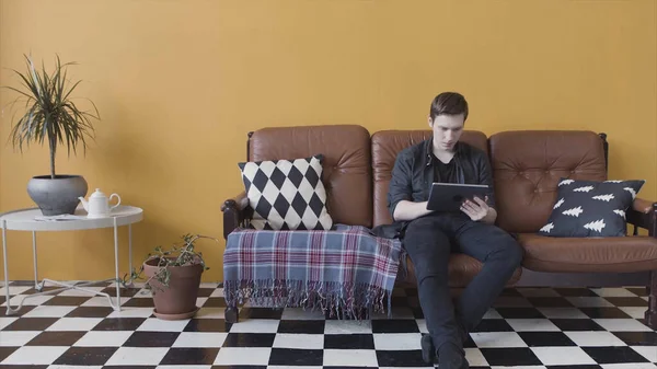 Guapo joven casual usando una tableta en casa sentado en un sofá en su sala de estar, navegando por Internet. Imágenes de archivo. Hombre serio utilizando su dispositivo en la habitación inusual brillante . — Foto de Stock