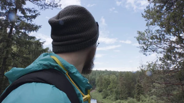 Närbild av hipster vandrare observera den gröna dalen på blå molnig himmel bakgrund. Lagerbilder. En skäggig man med runda glasögon med utsikt över en vacker skog. — Stockfoto