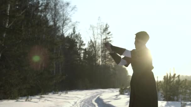 国服の剣を保持する若い男は、雪の森の前に立っている。行動だ男は、明るい冬の夕日に対して立って、セイバーを振ります. — ストック動画