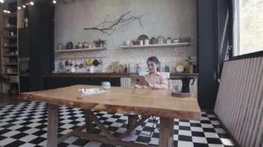 Teknoloji, internet ve ev konsepti, kadın tabletini kullanırken şık mutfaktaki ahşap güzel masada oturuyor. Stok görüntüleri. Cihazı evde olan kız.