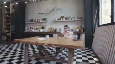 Teknoloji, internet ve ev konsepti, kadın tabletini kullanırken şık mutfaktaki ahşap güzel masada oturuyor. Stok görüntüleri. Cihazı evde olan kız.