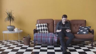 Evde tablet kullanan yakışıklı genç adam oturma odasındaki kanepede oturup interneti karıştırıyor. Stok görüntüleri. Ciddi bir adam cihazını alışılmadık bir odada kullanıyor..