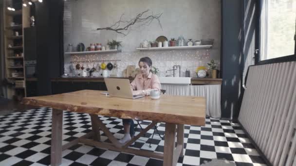 Žena pracující doma na notebooku ve stylové a prostorné moderní kuchyni, koncept na volné noze. Záběry ze skladu. Mladá krásná žena sedící u stolu s šálkem horkého nápoje a notebookem. — Stock video