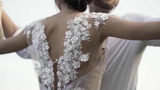 Rückansicht eines wunderschönen weißen Brautkleides einer jungen schlanken Braut mit sanften Blumen und Rückenausschnitt. Aktion. Liebevolle ang Umarmung Braut und Bräutigam auf hellem Himmel Hintergrund. — Stockvideo
