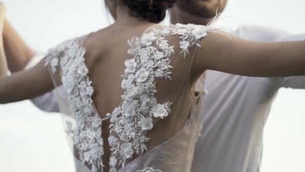 Vista posteriore di un bellissimo abito da sposa bianco di una giovane sposa magra con fiori delicati e ritaglio indietro. Azione. Amare ang abbracciare sposa e sposo su sfondo cielo luminoso . — Video Stock