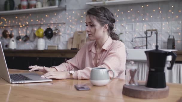 A young woman sitting at the kitchen table using a laptop. Stock footage. Beautiful brunette working at home on her computer with a cup of hot drink standing on the wooden table. — Stock Video